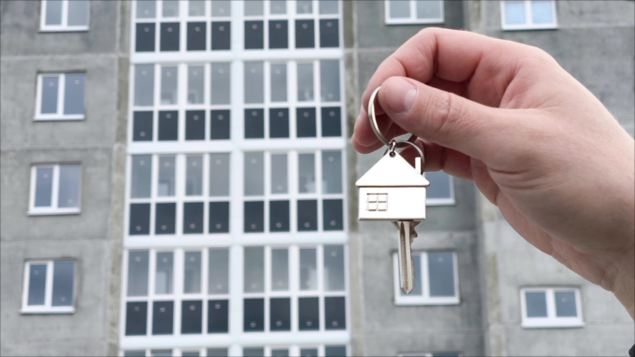 Hand hält Hausschlüssel mit Schlüsselanhänger in Form eines Hauses vor unsaniertem Mehrfamilienhaus – Symbol für Eigentümerwechsel und Sanierungspflichten laut Gebäudeenergiegesetz.